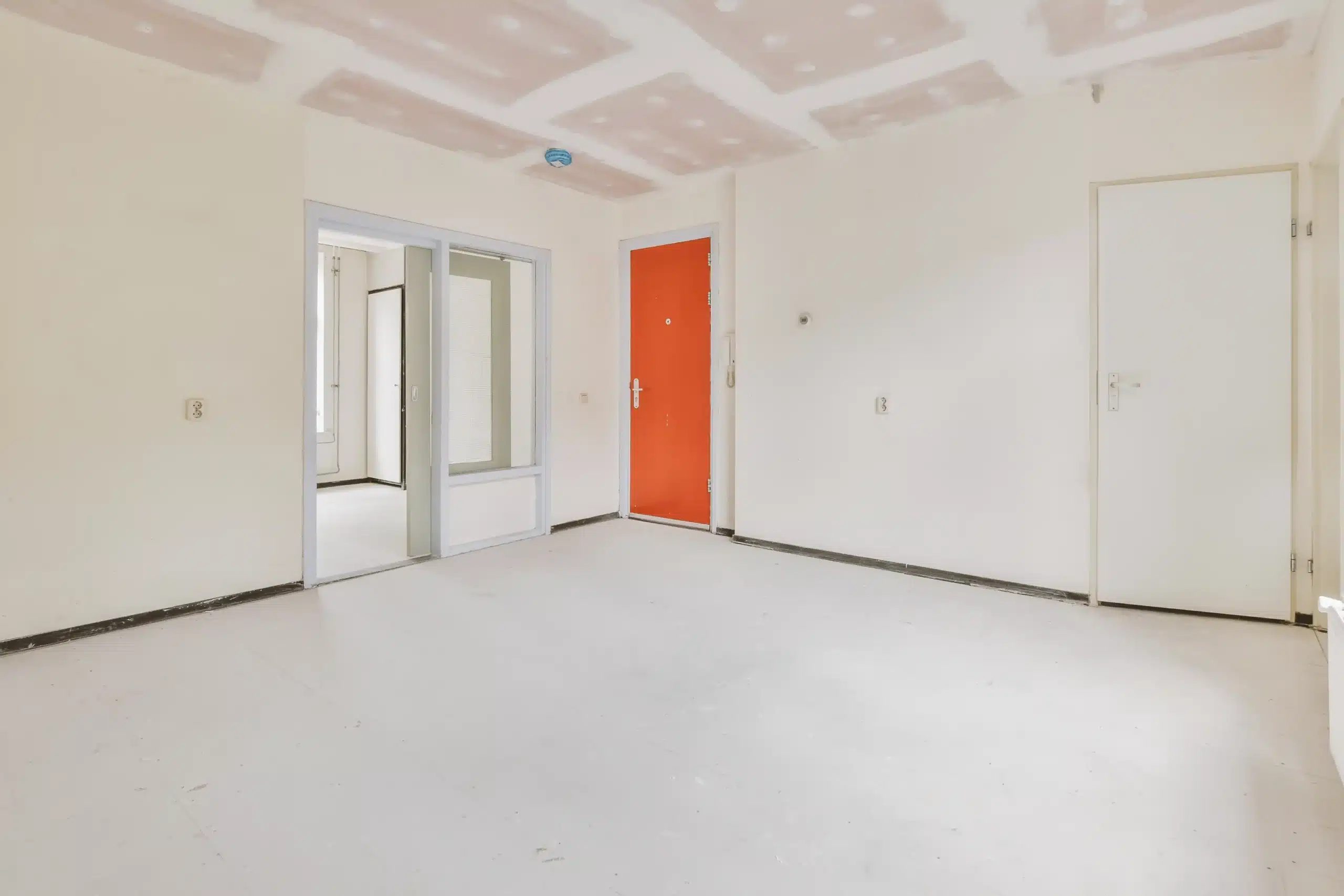 Empty room under renovation with white walls, unfinished ceiling, and a bright orange door